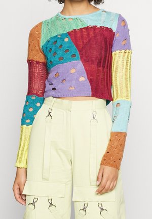 Woman wearing a multicolored patchwork knit cropped sweater with holes and light beige cargo pants featuring multiple silver clasps.