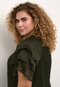 Blouse plissée vert foncé avec des manches à volants superposés. Texture transparente avec des rayures verticales ; encolure ronde et coupe décontractée.