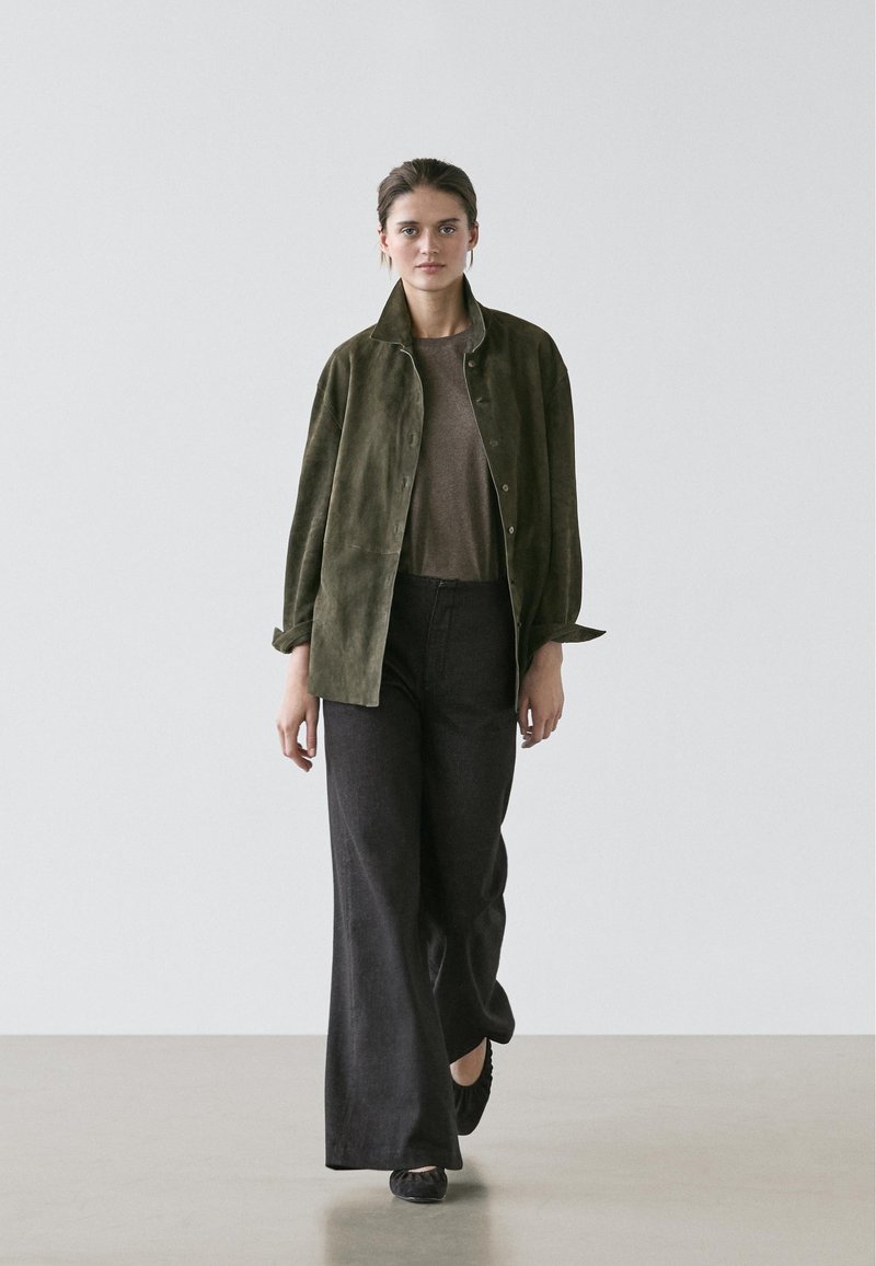 Veste en daim vert olive avec col, haut gris, pantalon large noir et ballerines noires, sur fond uni.