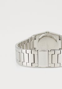 Silverfärgad klocka i rostfritt stål med länkarmband, oktagonformad klockkans, och en slät yta som visar minimal branding på baksidan.