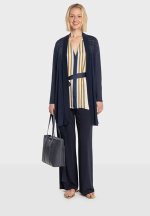 Tmavomodrý otvorený kardigán cez pruhovanú blúzku s žltými a bielymi pruhmi, kombinovaný s tmavými širokými nohavicami a plochými sandálmi. Čierna taška na rameno.