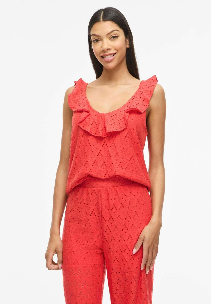 Femme souriante portant un haut rouge sans manches avec un col à volants et un pantalon texturé assorti, debout dans une posture détendue sur fond blanc.