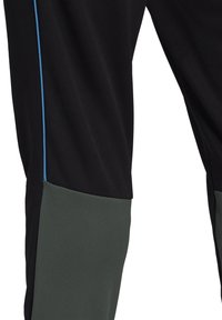 Gros plan sur un pantalon noir avec des poignets gris et une fine bande verticale bleue le long de la couture latérale sur la cuisse.