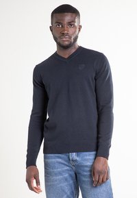 Maglione a V blu navy in tessuto a costine con maniche lunghe, caratterizzato da un piccolo logo sul petto e una fascia aderente in vita.