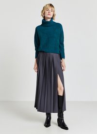 Türkisfarbener Strickpullover mit hohem, umgeschlagenem Kragen, kombiniert mit einem gefältelten grauen Rock mit seitlichem Schlitz. Schwarze Knöchelschuhen runden das Outfit ab.