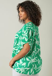 Top verde con un corte suelto, mangas cortas y un patrón floral blanco. Hecho de una tela liviana con un acabado texturizado.