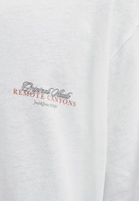 Balts kokvilnas T-krekls ar drukātu dizainu, kurā rakstīts "Original Studio" melnā krāsā un "REMOTE CANYONS" sarkanā krāsā. Mīksta tekstūra, ikdienišķs piegriezums.