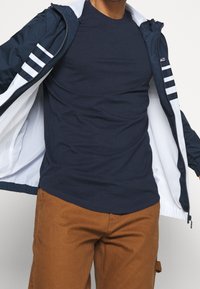 Veste zippée marine avec des accents de rayures blanches, portée sur un t-shirt marine et un pantalon marron. La veste possède une doublure en maille et des poignets élastiques.