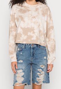 Person i beige tie-dye långärmad skjorta och högmidjade slitna blå denimshorts som står mot en neutral bakgrund.