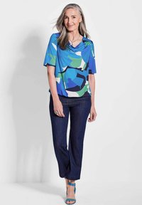 Blaues kurzärmliges Oberteil mit abstrakten grünen und marineblauen Mustern, kombiniert mit dunkelblauen Jeans und blauen Absatzsandalen. Glatter Stoff, lockere Passform.
