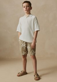 Witte korte mouw polo, beige korte broek met witte palmboomprint en bruine sandalen. Gladde textuur en losse pasvorm.