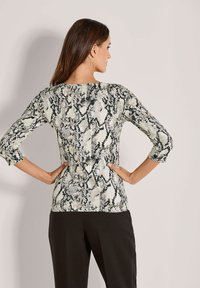 Frau mit langen braunen Haaren, die ein enges, langärmliges Oberteil mit Schlangenprint und schwarze Hosen trägt, steht mit den Händen in den Hüften und blickt weg.