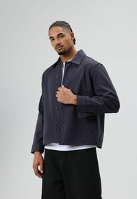 Donkerblauwe jas met knopen, wijde kraag, lange mouwen en een losse pasvorm. Draagt over een wit T-shirt en gecombineerd met een zwarte broek.