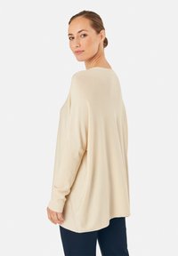 Beiger, oversized Pullover mit gerippter Textur, weiten Ärmeln und abgerundetem Saum, kombiniert mit marineblauen Hosen, seitlich gezeigt.