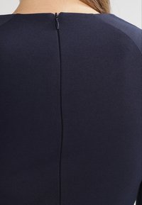 Dunkelblaues Kleid aus glattem Stoff mit einem zentralen Reißverschluss am Rücken und dezenten Nahtdetails an den Schultern.