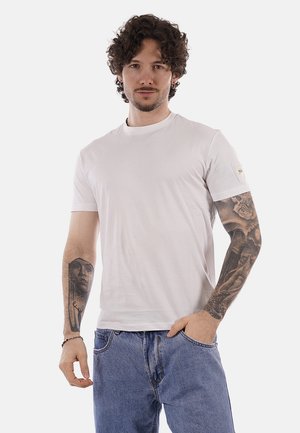 Uomo con capelli ricci che indossa una t-shirt bianca semplice e jeans blu, mostrando tatuaggi dettagliati su entrambe le braccia, in piedi con una mano in tasca.