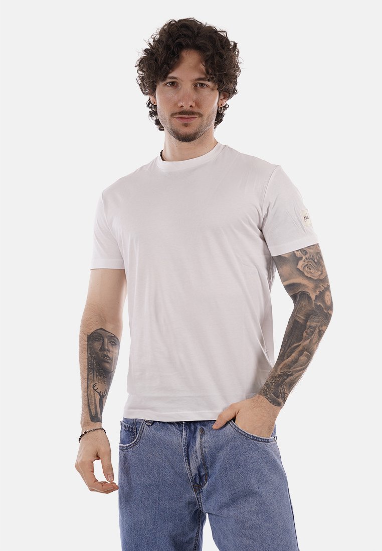 Homme aux cheveux bouclés portant un t-shirt blanc uni et un jean bleu, montrant des tatouages détaillés sur les deux bras, debout avec une main dans la poche.