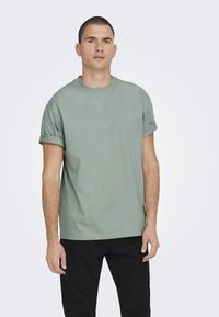Lichtgroene katoenen T-shirt met opgerolde korte mouwen, ontspannen pasvorm en ronde halslijn, gecombineerd met zwarte broek. Eenvoudig en onversierd ontwerp.