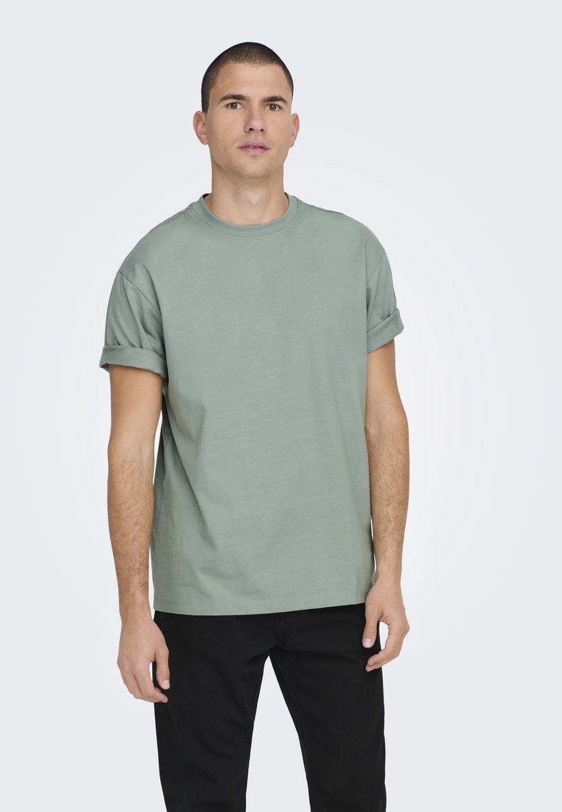 Lichtgroene katoenen T-shirt met opgerolde korte mouwen, ontspannen pasvorm en ronde halslijn, gecombineerd met zwarte broek. Eenvoudig en onversierd ontwerp.
