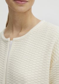 Cardigan w kolorze kremowym o fakturowanym materiale, z okrągłym dekoltem, z delikatnym geometrycznym wzorem i zapięciem z przodu.