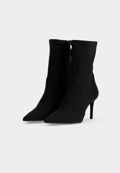 Paire de bottines noires à bout pointu et hauts talons stilettos, exposées sur un fond clair uni.
