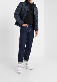 Puffetjakke i marineblå, sort hættejakke, mørke denimjeans og hvide sneakers. Har quiltet tekstur og opsmøg på jeansene.
