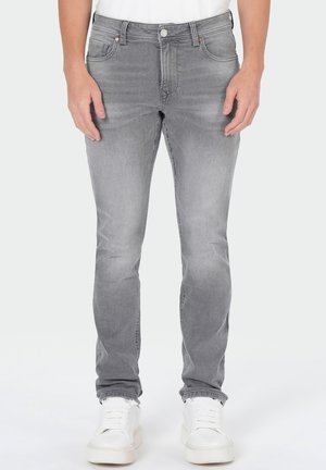 Mann trägt eng geschnittene graue Jeans und weiße Sneaker, steht vor schlichtem Hintergrund, Hände liegen seitlich am Körper.