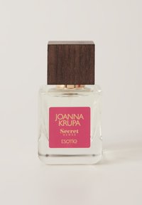 Przezroczysta szklana butelka perfum z kwadratową ciemnobronzową nakrętką. Zawiera różową etykietę ze złotym tekstem, przedstawiającą nazwę marki oraz nazwę zapachu.