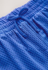 Modré krátke nohavice s potlačou a textúrovaným sieťovým dizajnom. Majú elastický pás a zodpovedajúce šnúrky na stiahnutie pre úpravu veľkosti.