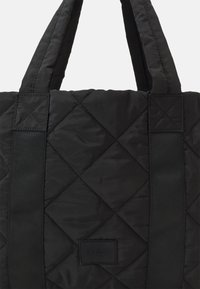 Borsa a spalla nera trapuntata realizzata in tessuto liscio e imbottito, con ampie cinghie in tessuto e una toppa con logo rettangolare cucita su un lato.