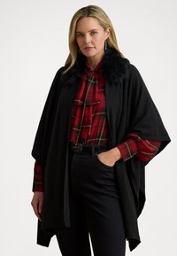 Poncho negru cu guler din blană artificială, purtat peste o bluză cu carouri roșii. Are un design fluid, mâneci lungi și o croială lejeră.