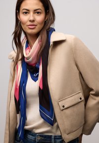 Beige fleecejakke med lynlås, der har en krave og en lomme med trykknap. Multifarvet silkeskærf med geometriske mønstre og blå accenter.