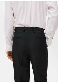 Zwarte op maat gemaakte broek met een gladde textuur, twee achterzakken en een vlakke voorkant. Draagt een lichtgestreept shirt.