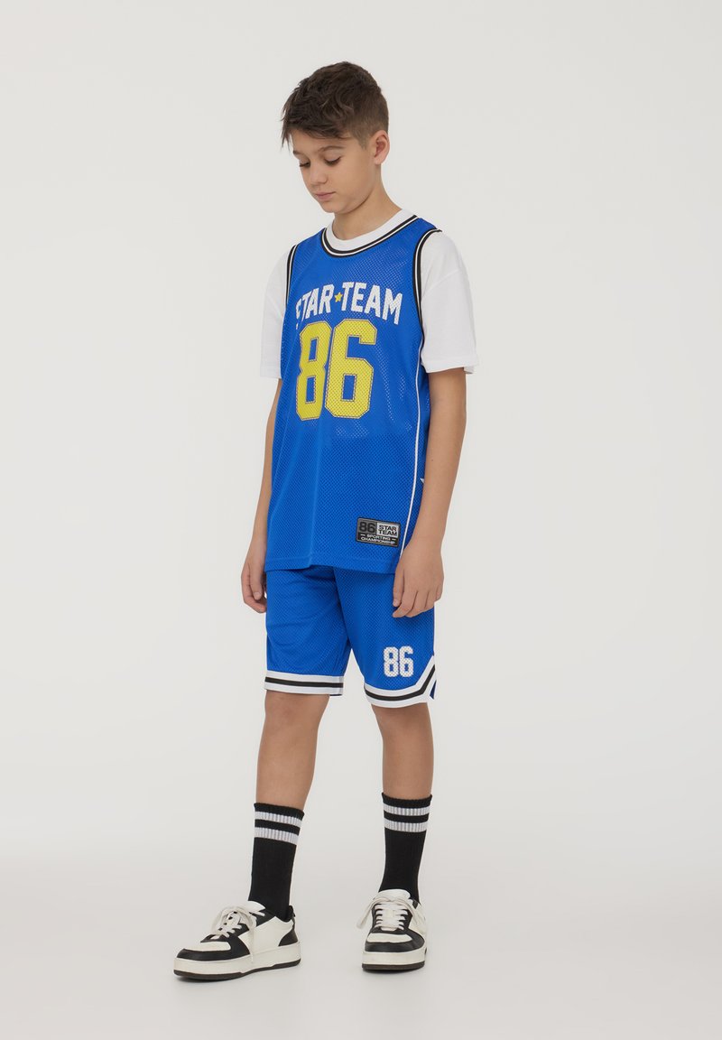 Maglia da basket blu con "STAR TEAM 86" in giallo, abbinata a pantaloni corti coordinati. Presenta dettagli a strisce bianche e nere. T-shirt bianca sotto.