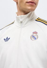 Άνδρας που φοράει λευκό μπουφάν με φερμουάρ με κίτρινες και ναυτικές ρίγες στους ώμους, λογότυπο Adidas στο αριστερό στήθος, σήμα της Ρεάλ Μαδρίτης στο δεξί στήθος.