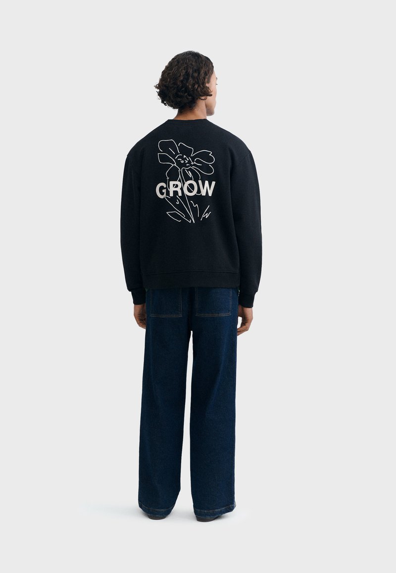 Personne aux cheveux courts et bouclés, portant un sweat-shirt noir avec une fleur blanche et le mot "GROW" dans le dos, et un jean bleu foncé ample, debout.