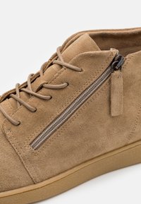 Baskets montantes en daim beige avec fermeture éclair latérale, lacets, col rembourré et semelle en caoutchouc, vues en gros plan.