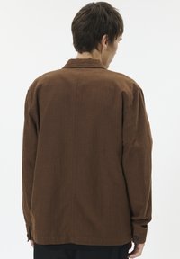 Muškarac viđen s leđa kako nosi labavu smeđu košulju s gumbima i tamne hlače na jednostavnoj bijeloj pozadini.