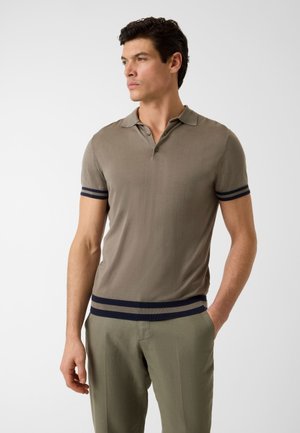 Mann trägt ein taupefarbenes Polohemd mit kurzen Ärmeln, navyblaue Streifen an Ärmeln und Saum, kombiniert mit hellolivgrüner Hose, schaut zur Seite.