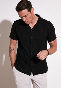 Camisa de botón negra de manga corta con cuello, de tejido texturizado y con dobladillo curvado, combinada con pantalones claros.