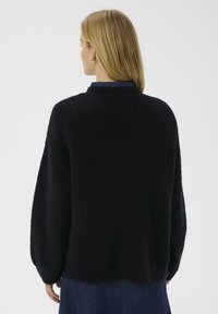 Schwarzer, flauschiger Strickpullover mit rundem Halsausschnitt, lockerer Passform und geripptem Saum. Übereinander getragen mit einem dunkelblauen Shirt, das aus weichem, strukturiertem Garn besteht.