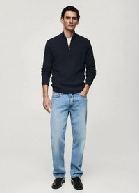 Navy knitted zip-up sweater, light blue straight-leg jeans, and black shoes. Model stands with hands in pockets, neutral background.