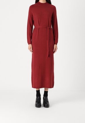 Kobieta ubrana w długą, sięgającą kostek czerwonym dzianinowym sukienkę z wiązaniem w talii, czarne skarpetki i czarne mokasyny, stoi na białym tle.