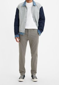 Veste matelassée grise et marine avec des accents en denim, associée à un pantalon gris clair et des baskets noires avec des semelles et lacets blancs.