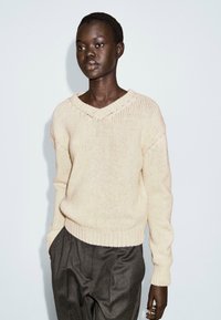 Pull en tricot beige clair avec un col en V, un ourlet côtelé et des manches longues ; associé à un pantalon noir texturé taille haute.