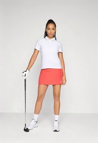 Hvit poloskjorte, rosa sportsskjørt, hvite sokker og joggesko. Holder en golfkølle. Nøytral bakgrunn, modellen står selvsikkert.