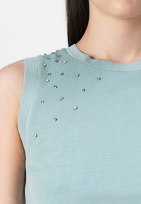 Mintgrønn ermeløs topp pyntet med sølvnitter på den ene skulderen, med en nærbildevisning av stoffet og halslinjen.