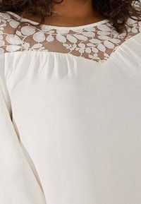 Vit blus med blommigt spets yfteil och V-formad halsringning, mjuk tygtextur och rynkade detaljer vid axlarna.