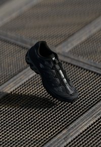 Black Salomon trail running shoe floating above metal grated surface with crossbeam pattern in soft light.