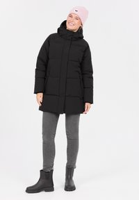 Sort puffersjakke med høj krave, horisontalt quiltet mønster, frontlommer med lynlås, parret med grå jeans og sorte gummistøvler til anklerne.